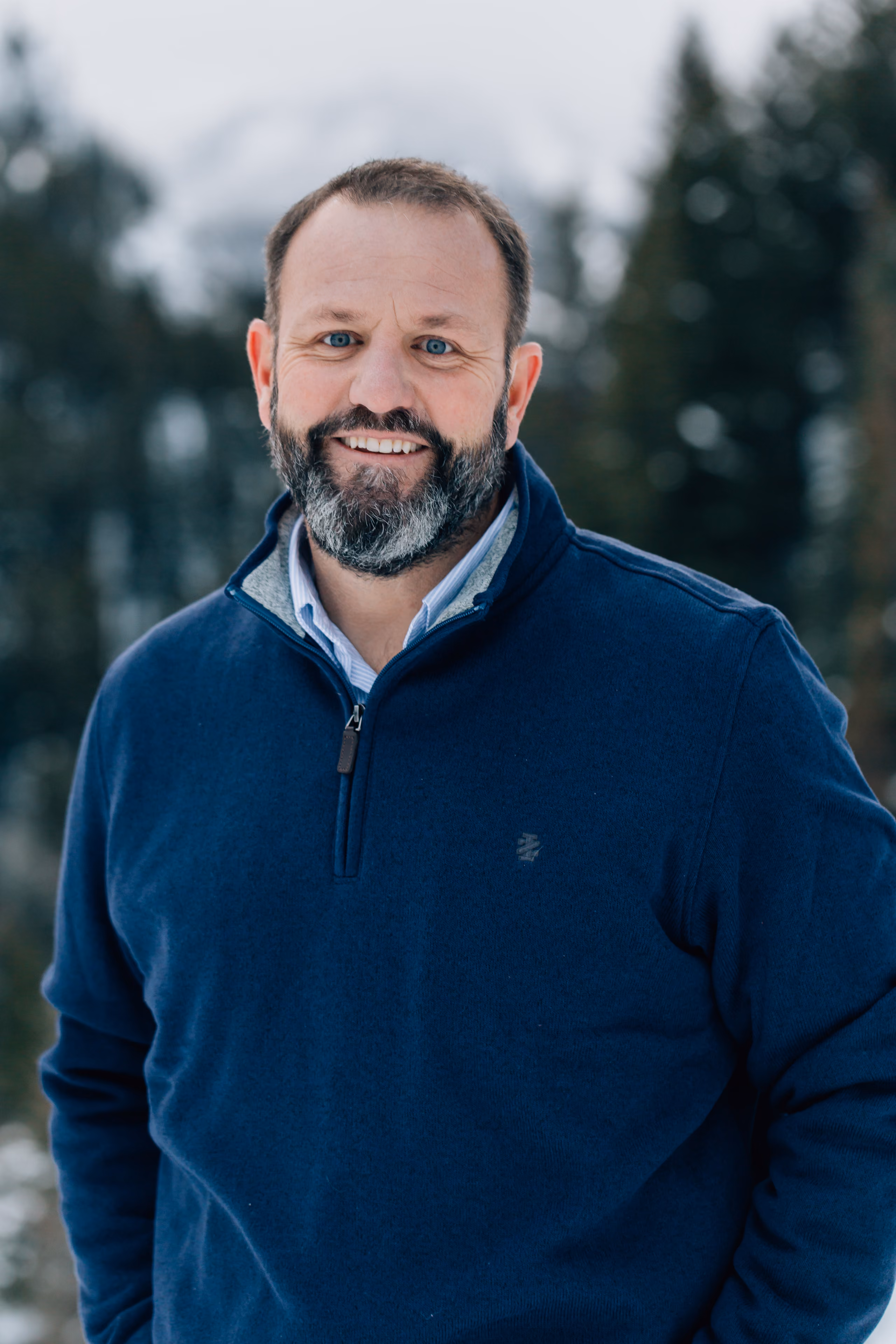Smiling bearded man in blue pullover outdoors with snowy trees and mountain in background.