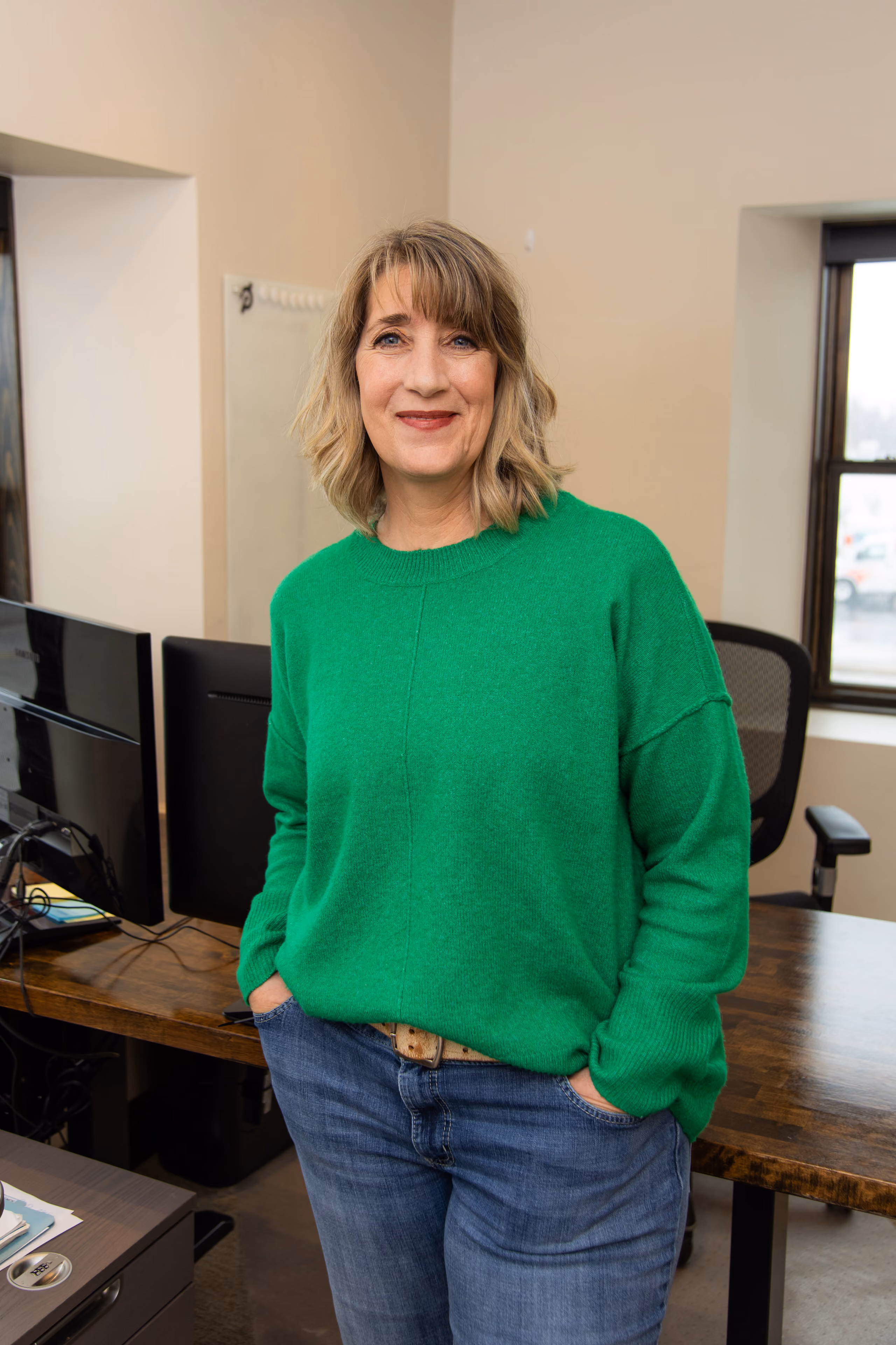 Smiling woman in green sweater and jeans standing in an office with computer monitors and wooden desk.