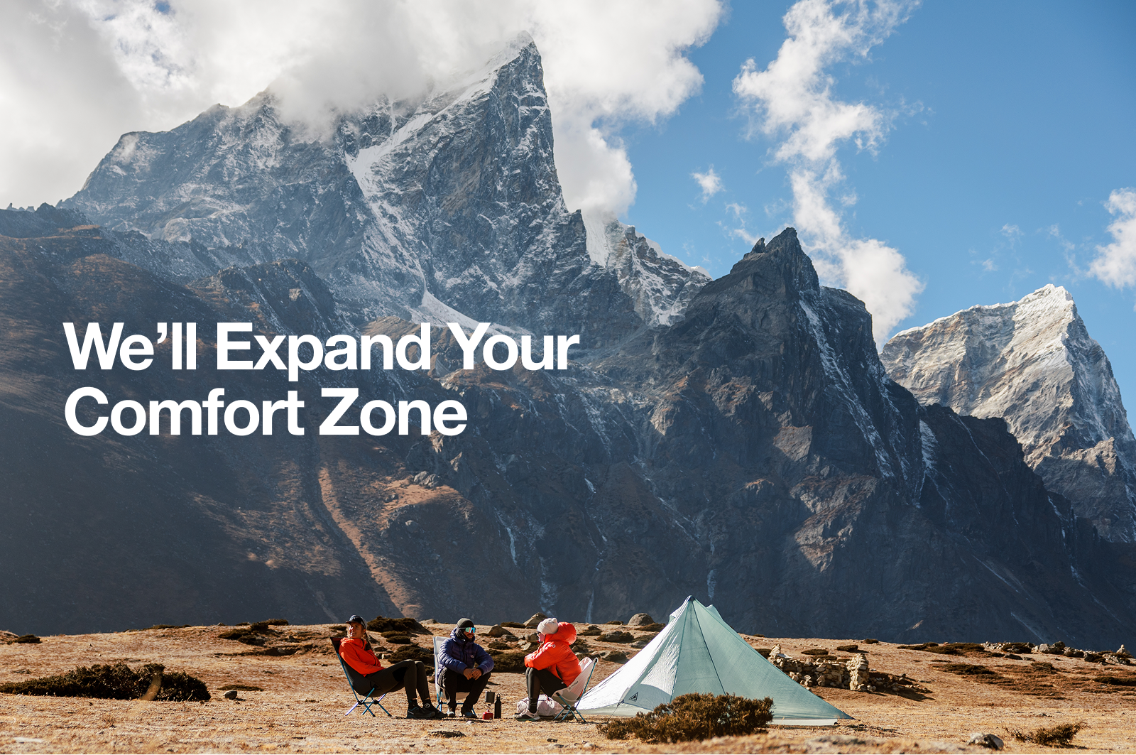 Three people sitting on camping chairs beside a light blue tent in a mountainous area under a partly cloudy sky.