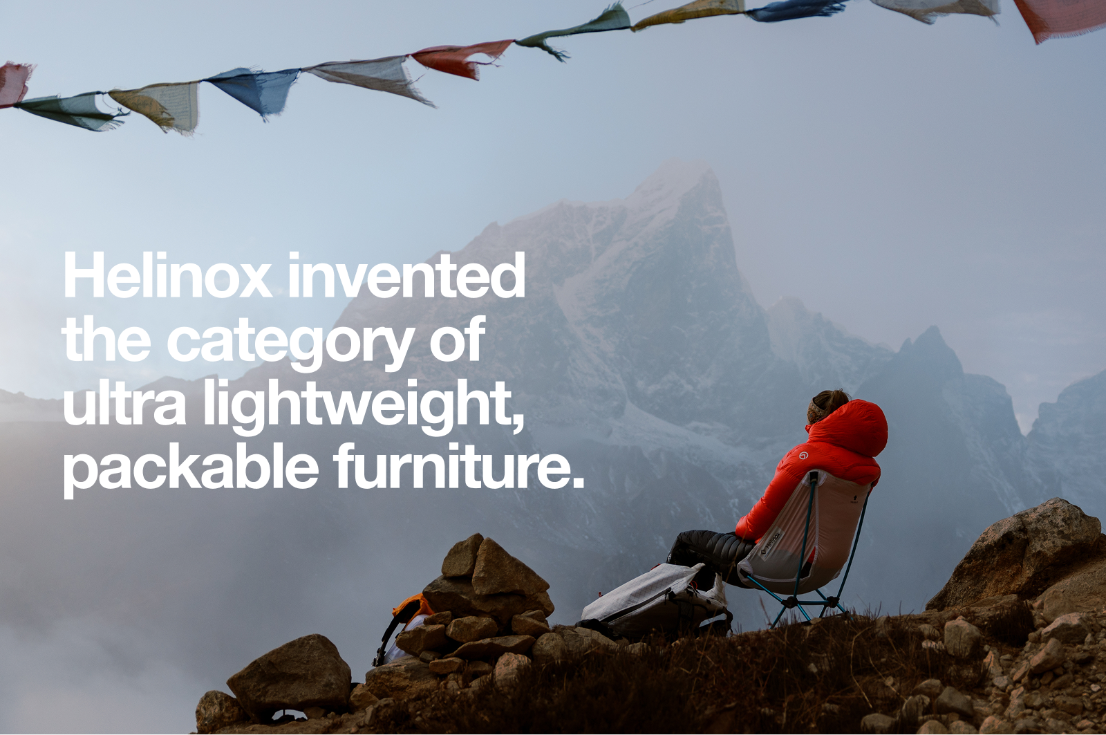Person in a red jacket sitting on a lightweight camping chair on rocky terrain, overlooking foggy mountains.
