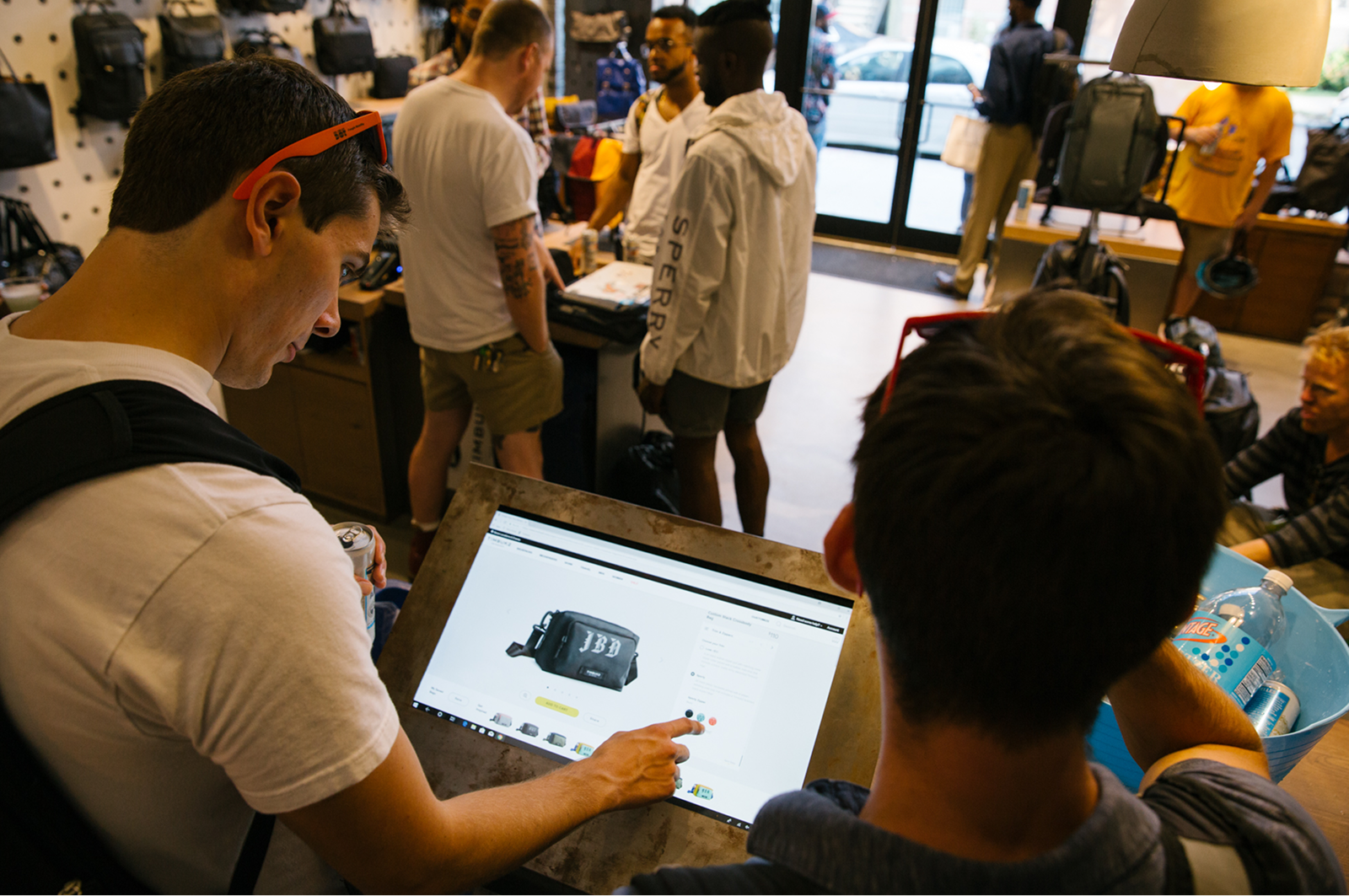 Two men looking at an online store page displaying a black bag on a touchscreen monitor inside a retail shop with other customers and backpacks in the background.