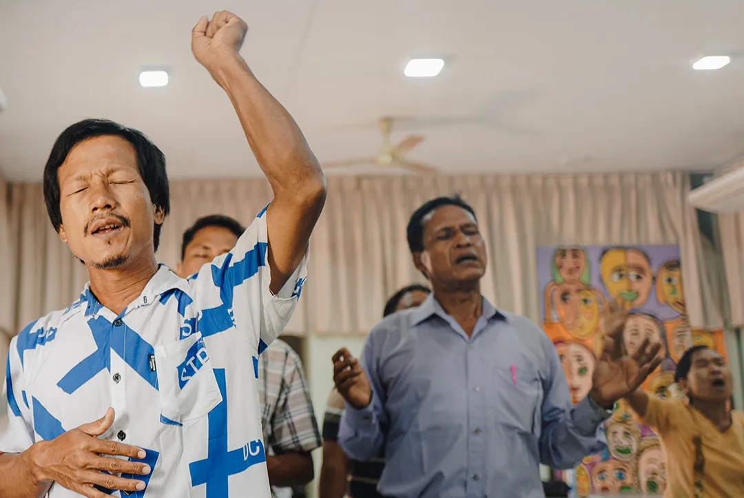 Group of men with closed eyes raising hands and singing or praying indoors.
