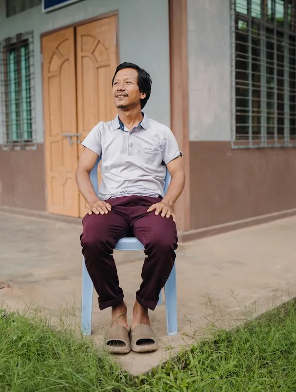 Man sitting on a plastic chair outside a building with wooden doors and barred windows, looking to his left.