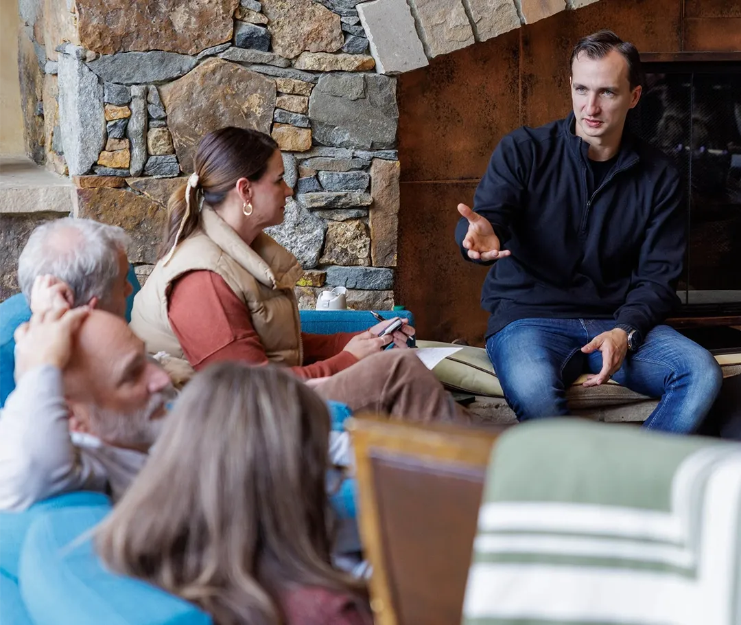 Man in black jacket speaking to a small group seated in a cozy room with a stone wall and fireplace.