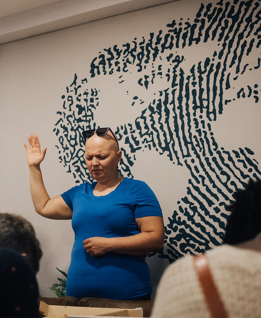 A bald woman in a blue shirt with sunglasses on her head raises her right hand and closes her eyes, standing in front of a wall with abstract dark blue patterns.