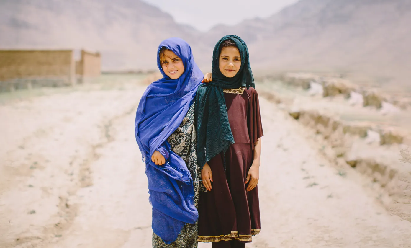 Two young girls wearing headscarves standing side by side on a dirt road with mountains in the background.