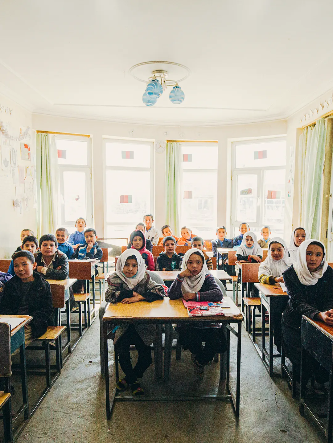 A classroom with children sitting at desks facing forward, some girls wearing white headscarves.