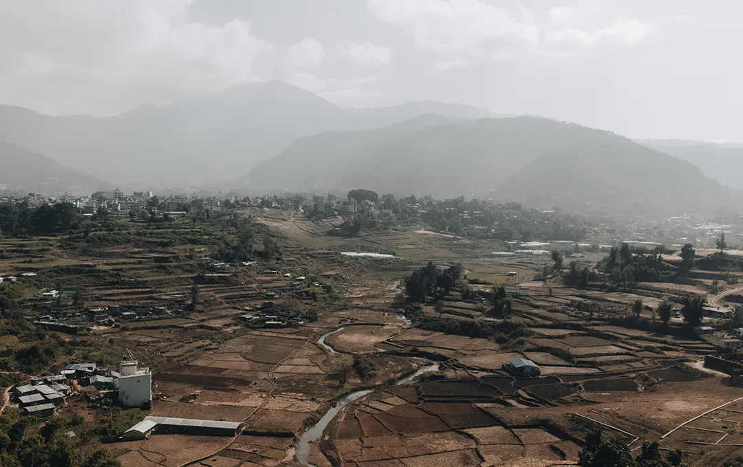 Aerial view of a rural landscape with terraced fields, a winding river, scattered houses, and mountains in the background under a cloudy sky.