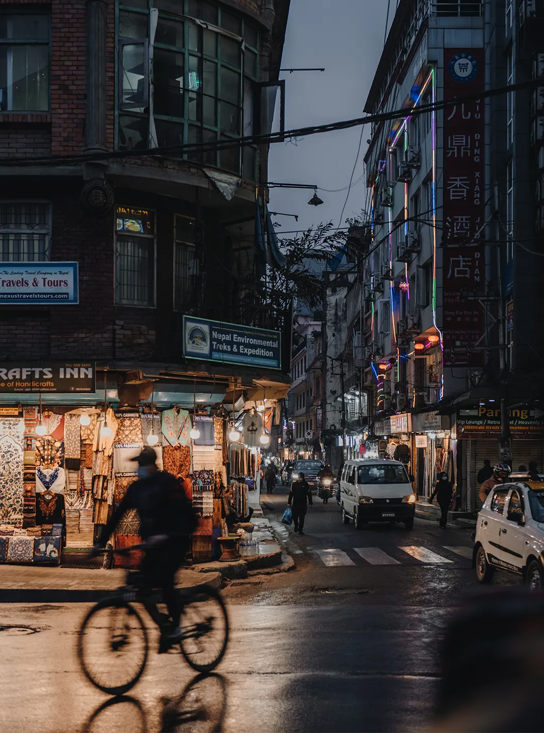 Evening street scene with a cyclist passing shops illuminated by warm lights and pedestrians walking in an urban area.