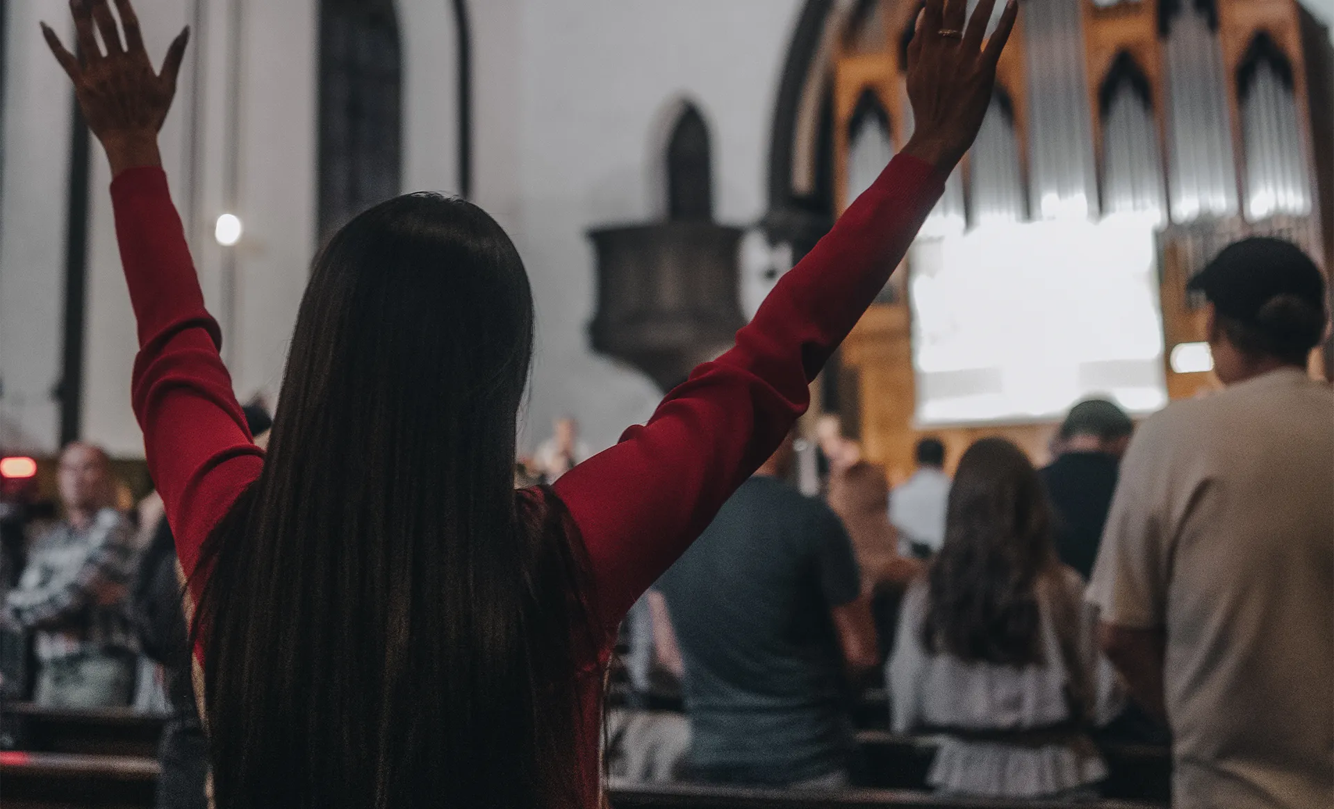 Person with long dark hair wearing a red sweater raising their hands in a church with a congregation in the background.