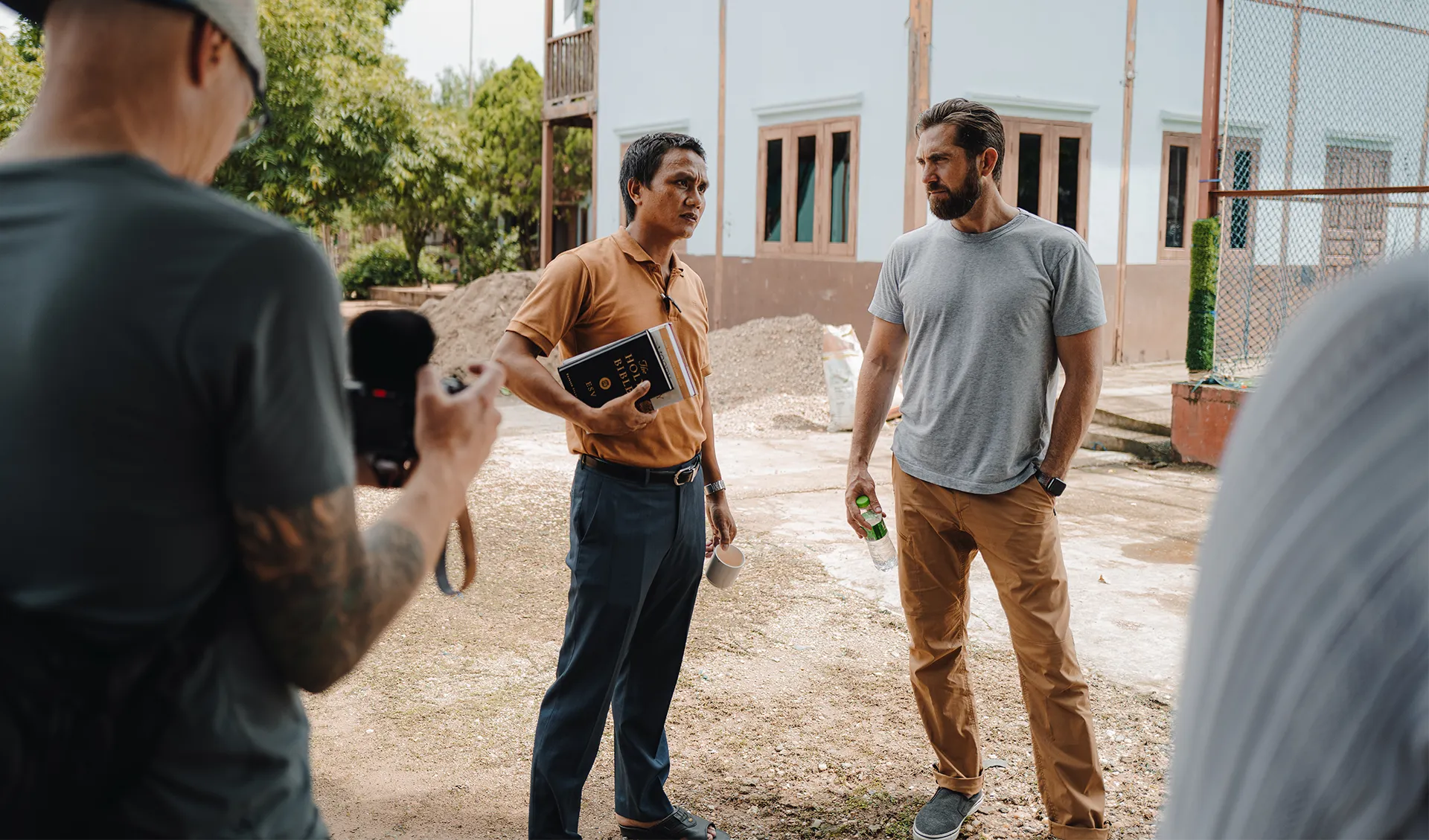 Two men standing outdoors engaged in conversation, one holding a Bible and a mug, the other holding a water bottle, with a third man in the foreground holding a camera.