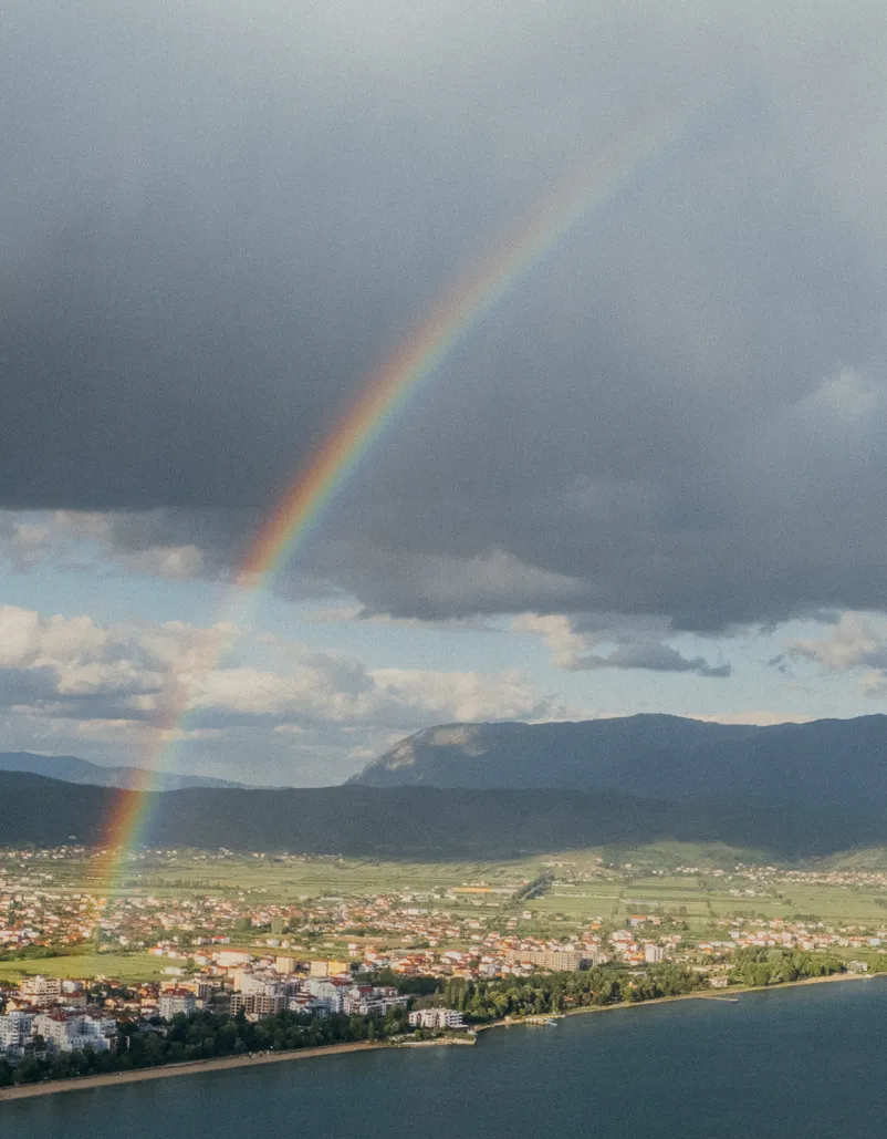 Rainbow arching over a coastal town with mountains and cloudy sky in the background.