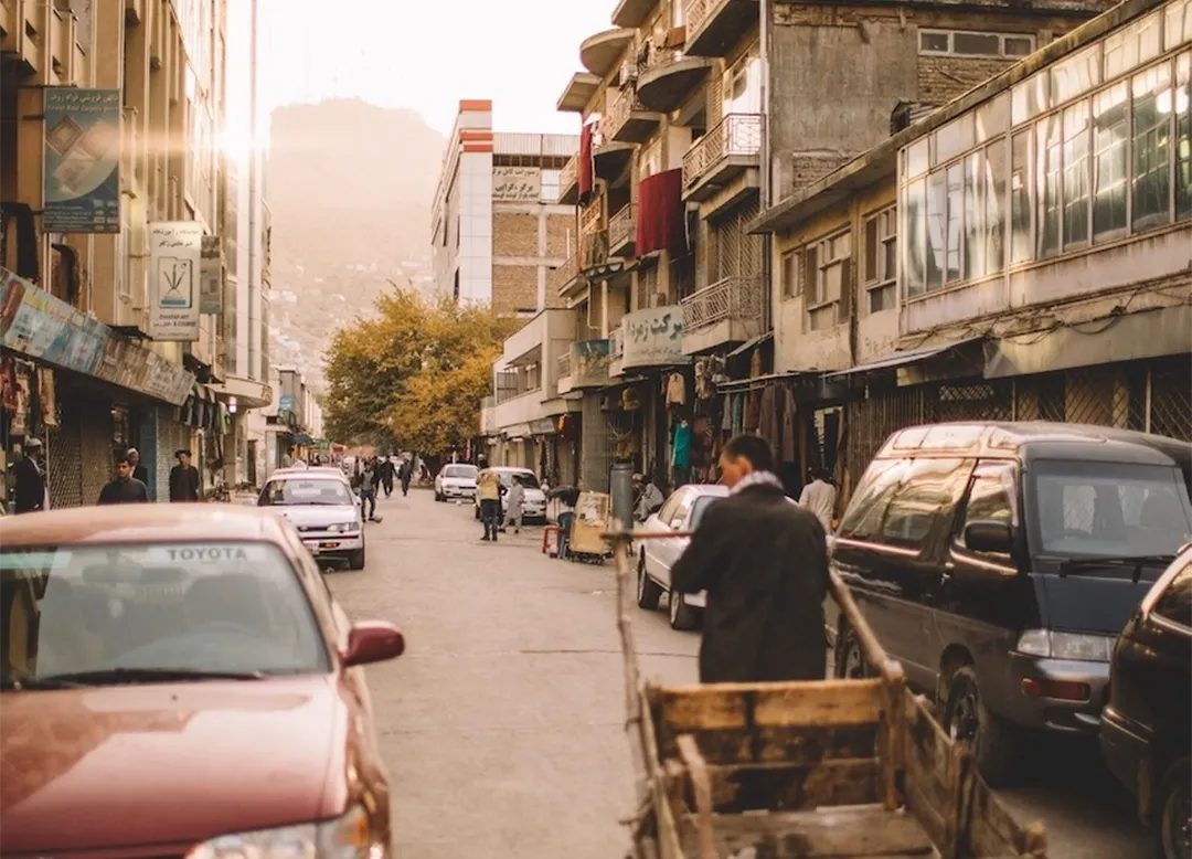 Street scene with people walking, parked cars, and a man pushing a wooden cart in an urban area with buildings on both sides.