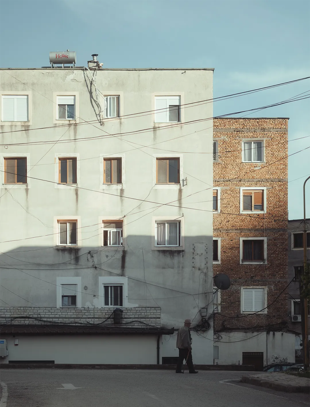 An elderly person walks with a cane on a street in front of adjacent beige and brick residential buildings under a clear blue sky.