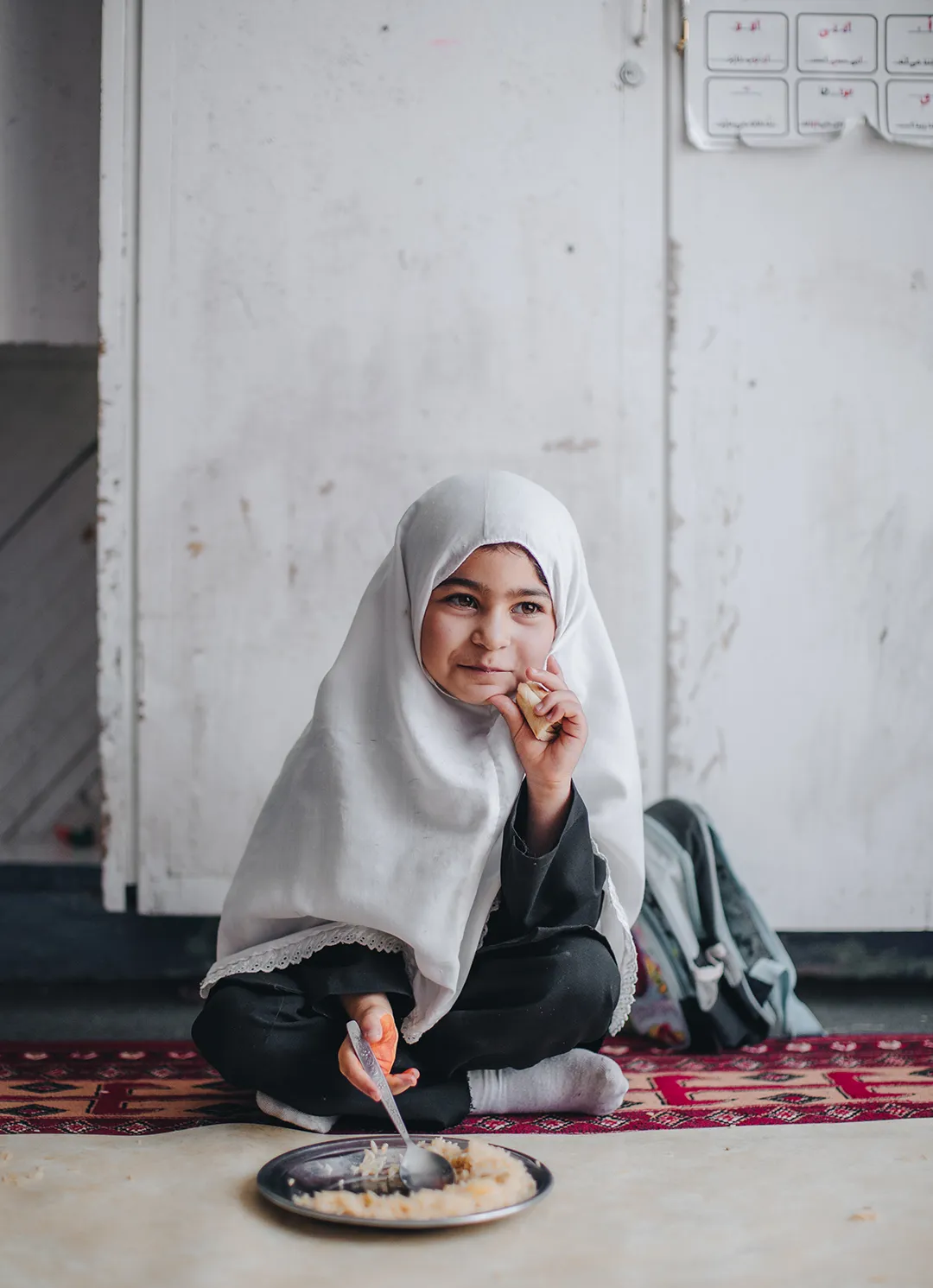 Young girl in a white hijab sitting cross-legged on a red patterned rug, eating food from a metal plate with a spoon and holding a piece of bread.