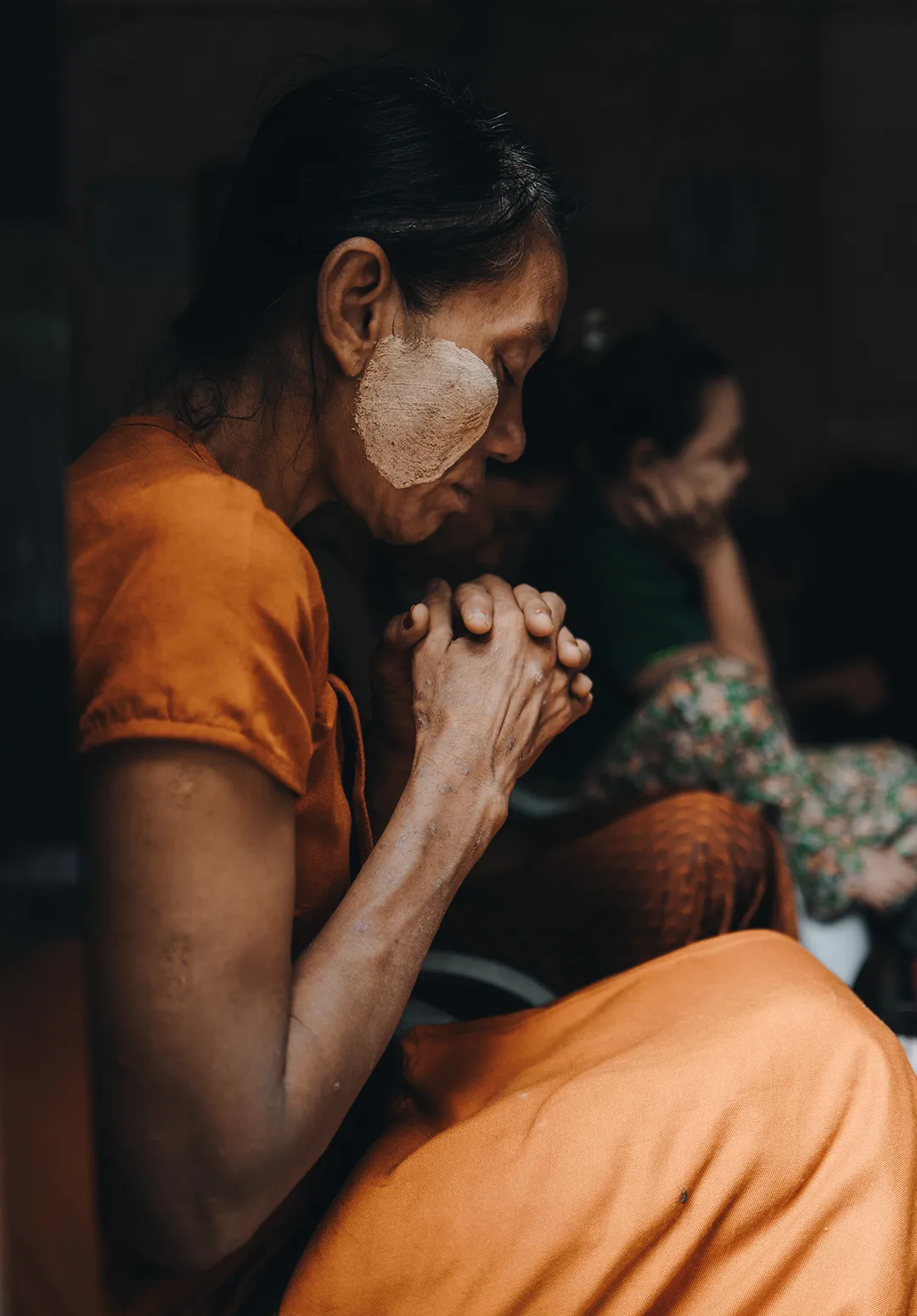 Side profile of a woman in orange clothing with thanaka paste on her cheek, sitting with eyes closed and hands clasped in prayer or contemplation.