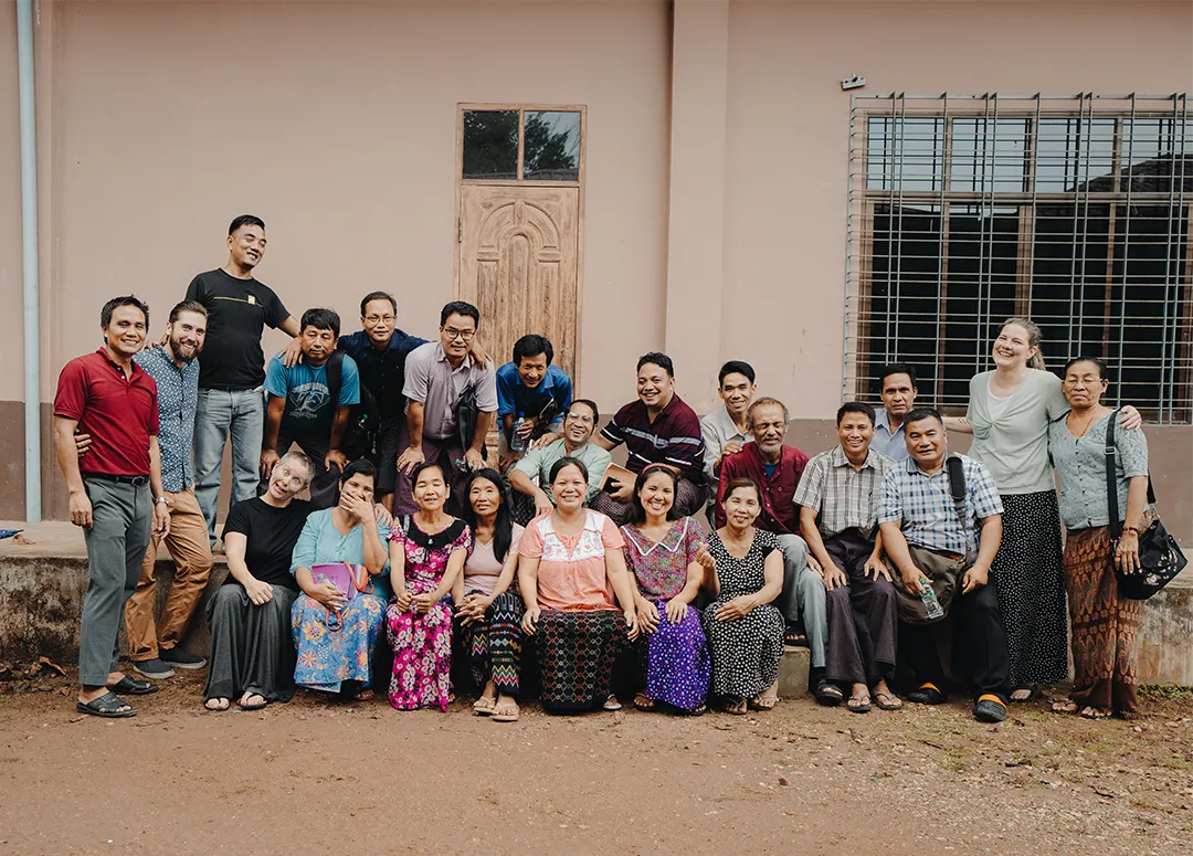 Group of diverse people smiling and posing for a photo outdoors in front of a beige building with a wooden door and window with metal bars.
