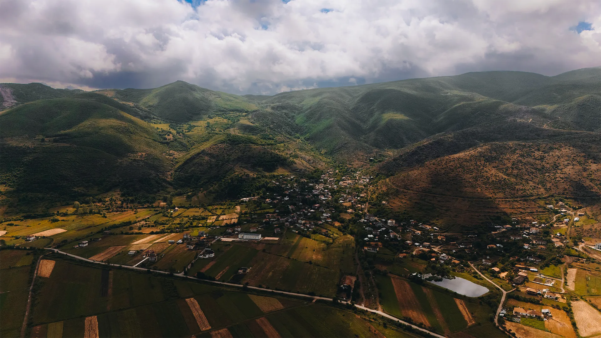 Aerial view of a rural village nestled in a valley surrounded by green and brown mountains under a cloudy sky.