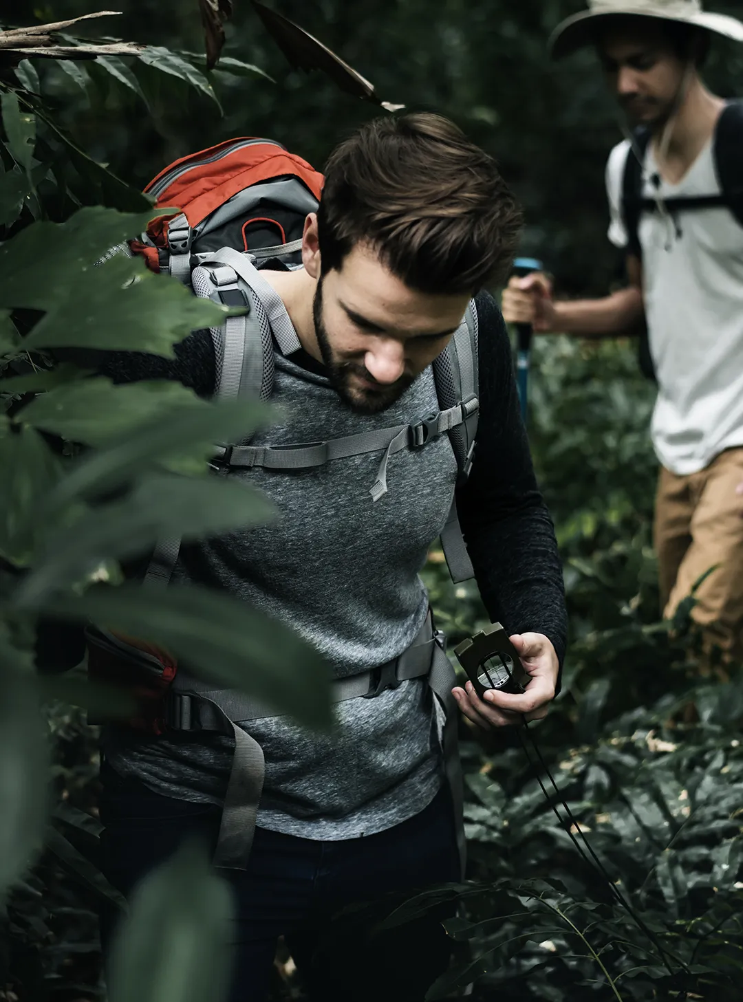 Two hikers in a dense forest with backpacks, one using a compass and the other holding a trekking pole.