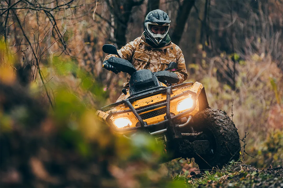 Person wearing a helmet and camouflage jacket riding a yellow ATV with headlights on through a muddy forest trail.