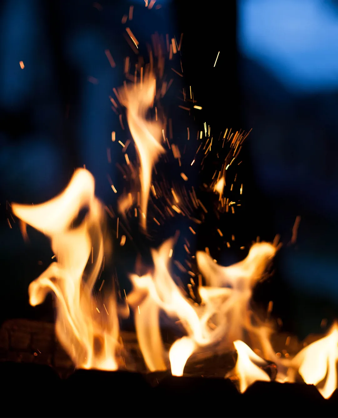 Bright orange flames and sparks rising from a burning fire with a dark blue blurred background.