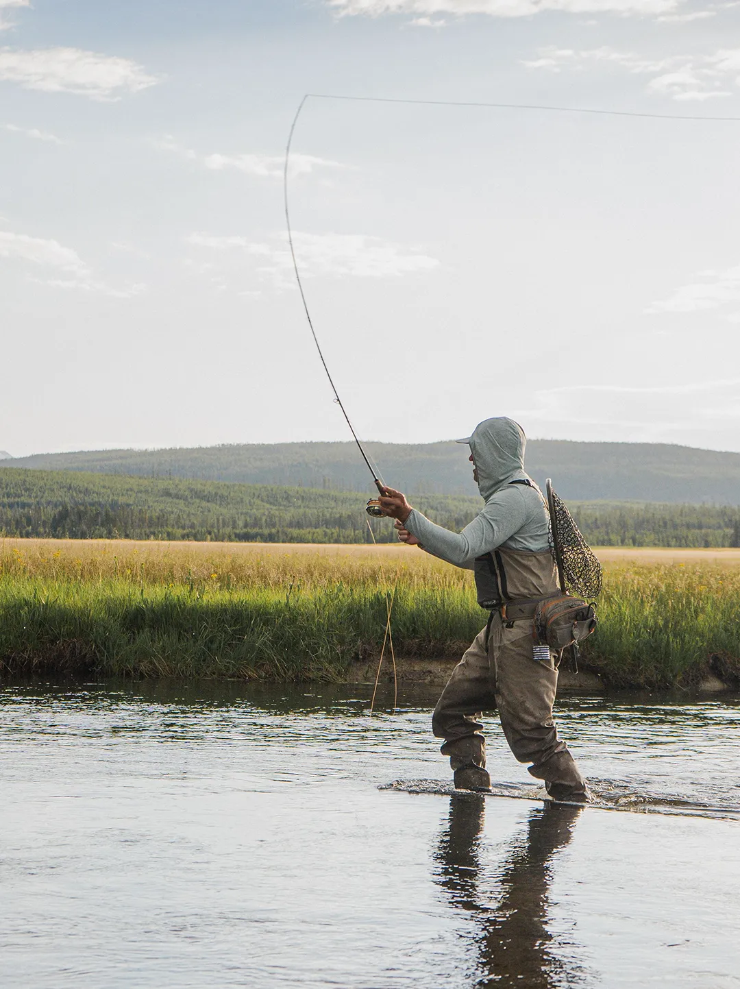 Person wearing a hooded jacket and waders fly fishing in a river with grassy banks and forested hills in the background.