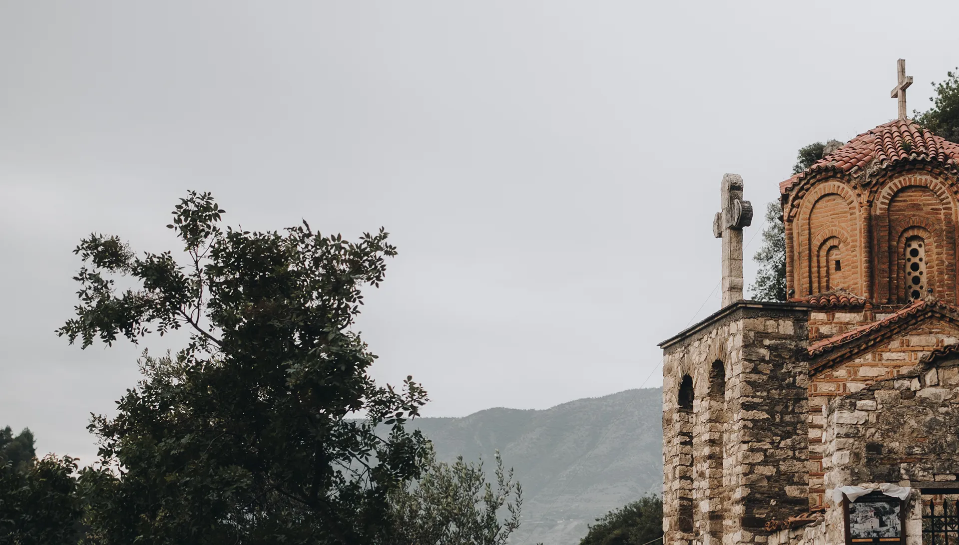 Stone and brick church with cross on rooftop and bell tower against a cloudy sky and mountainous background.