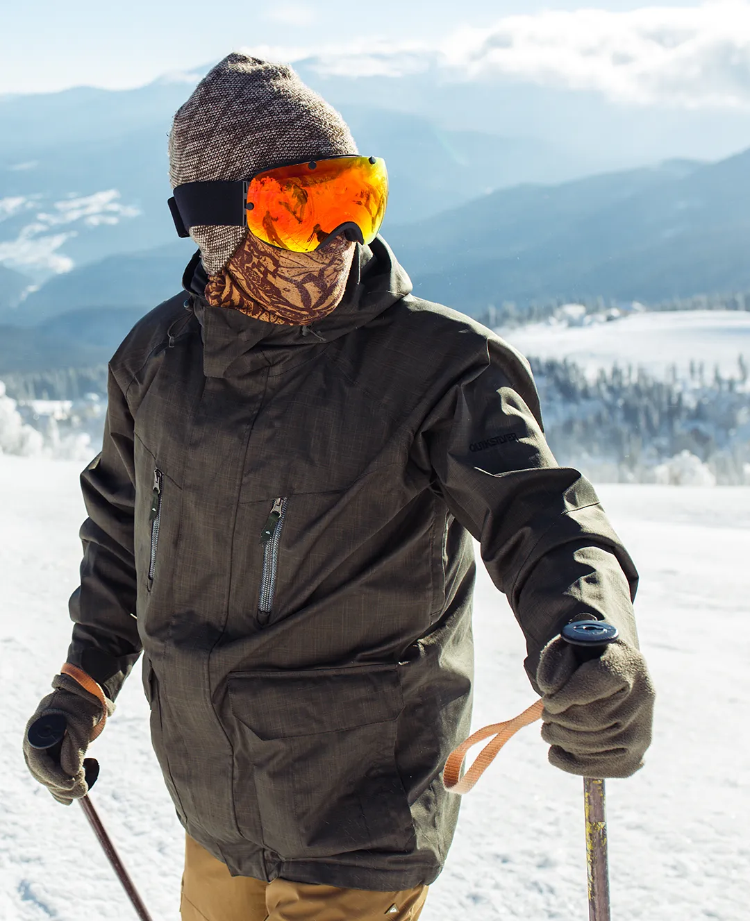 Person dressed in winter clothing with brown jacket, knit hat, orange ski goggles, and gloves holding ski poles on a snowy mountain.