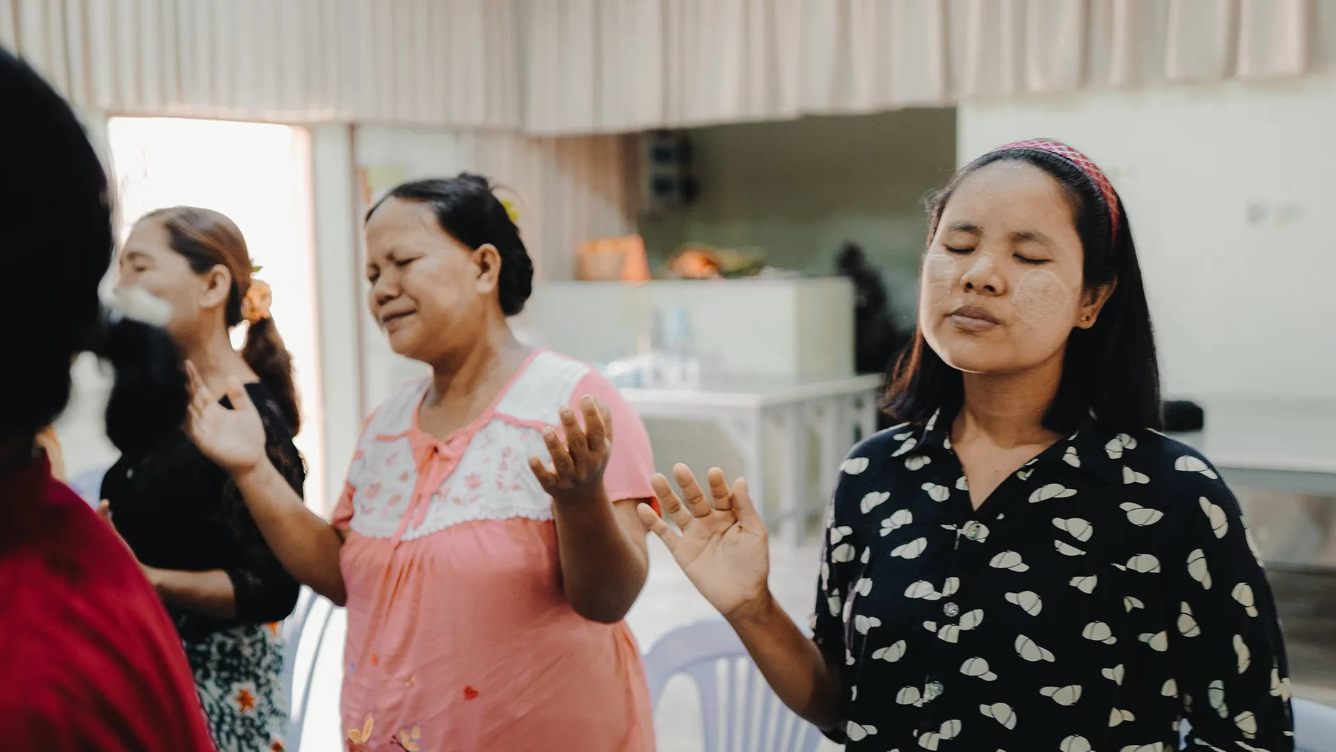 Three women standing indoors with eyes closed and hands raised, appearing to be in prayer or meditation.