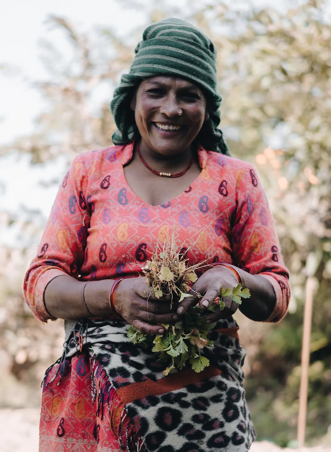 Smiling woman in a green striped headscarf and patterned red dress holding fresh herbs outdoors.