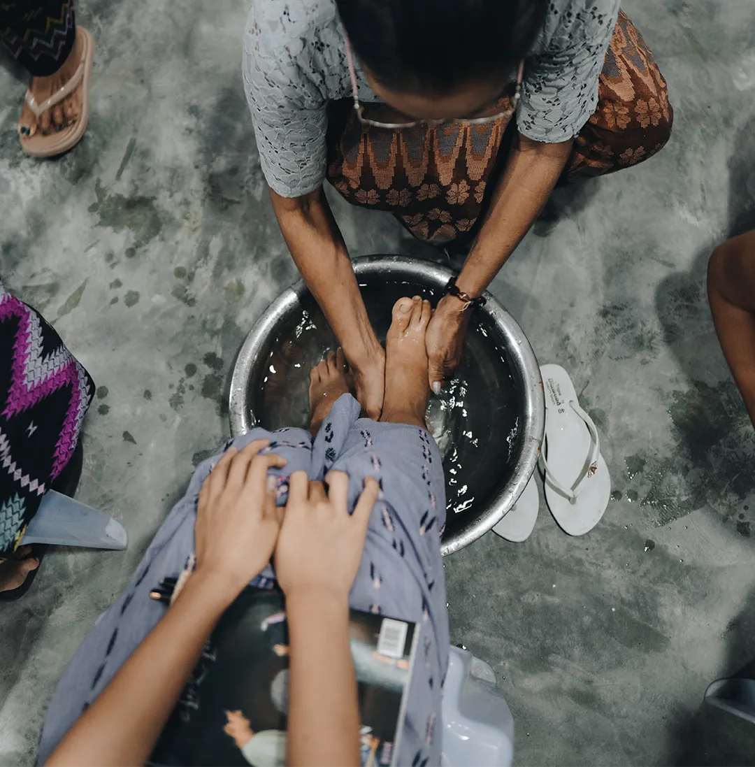 A person washing another’s feet in a metal basin filled with water on a concrete floor.