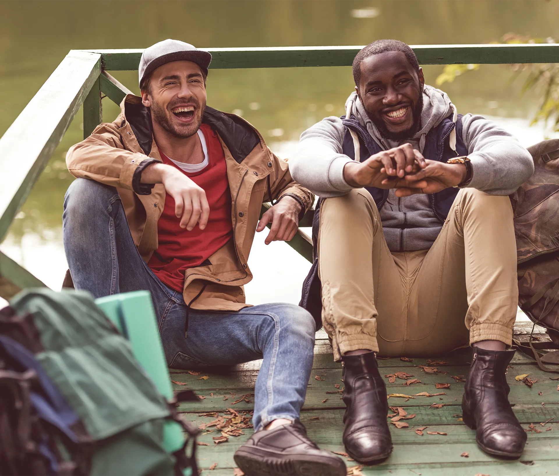 Two men sitting on a wooden dock by the water, smiling and laughing while dressed in casual outdoor clothing.