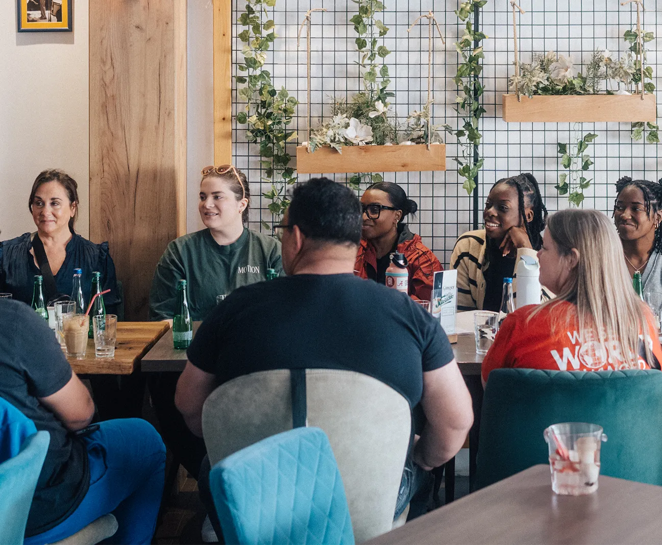 Group of people sitting at a table in a café with plants hanging on a grid wall behind them.