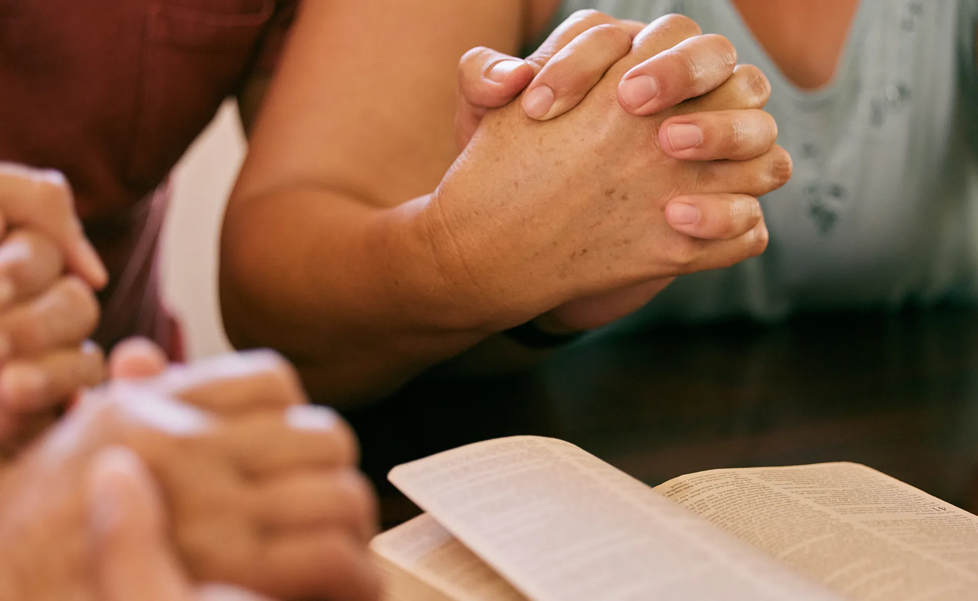 Two people holding hands in prayer over an open Bible on a table.