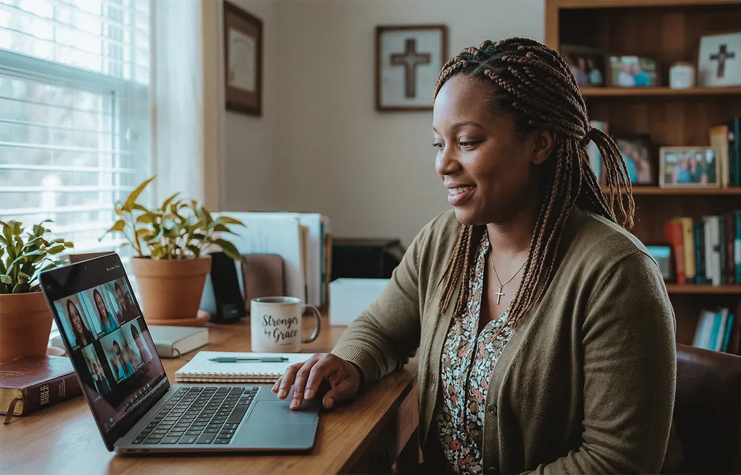 Smiling woman with braided hair and a cross necklace participating in a video call on a laptop at a wooden desk with plants, books, and a mug.