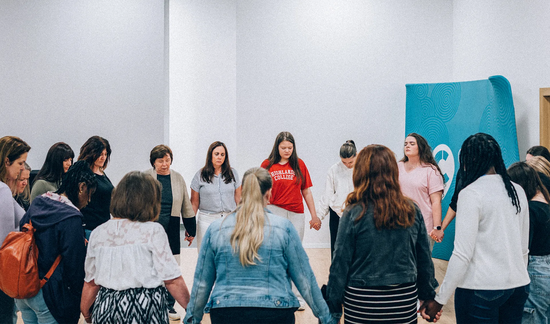 Group of women standing in a circle holding hands with eyes closed in a white room.