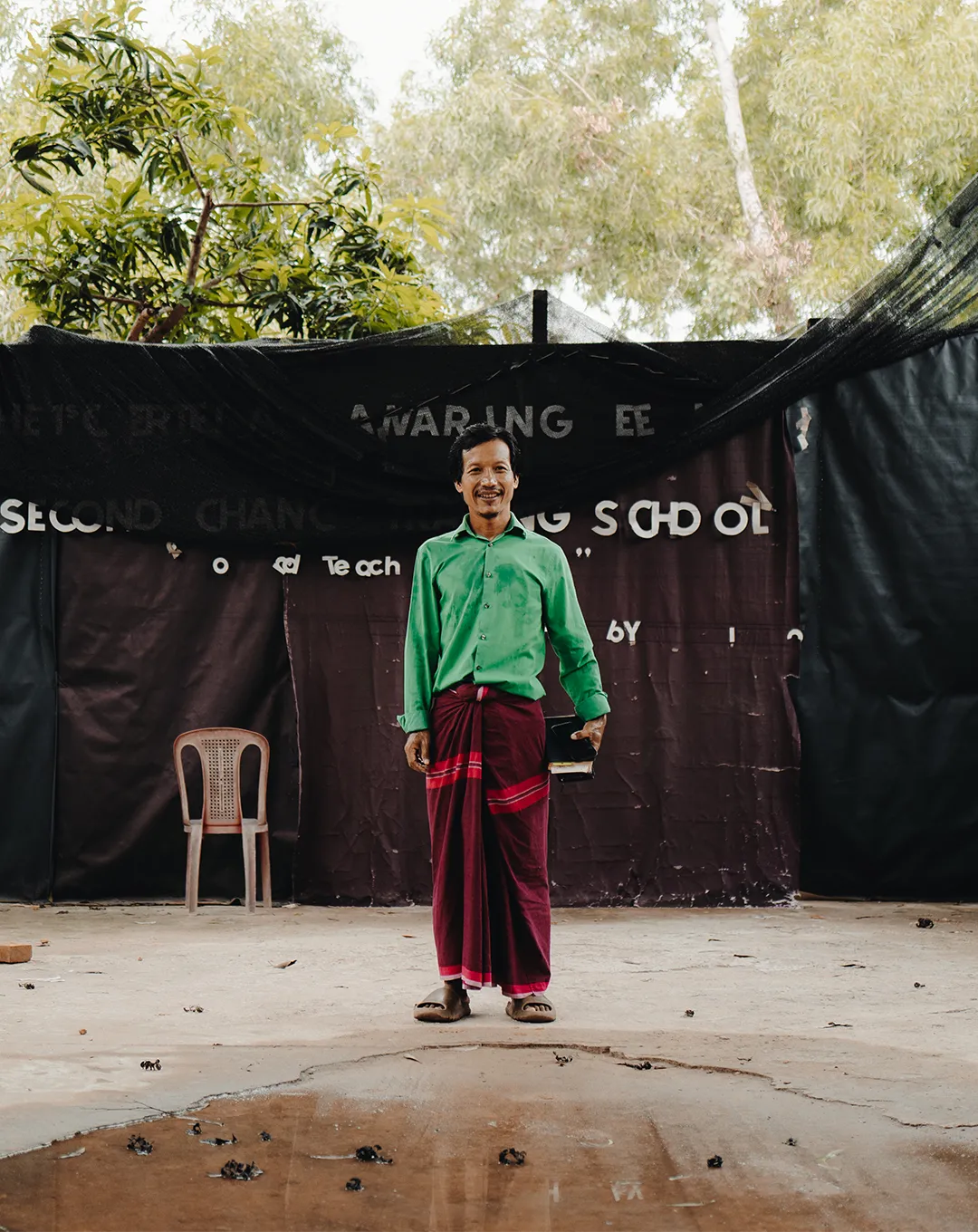 Man in a green shirt and maroon traditional wrap stands barefoot indoors holding books, with trees and a chair behind him.
