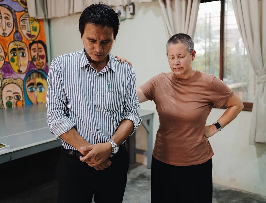 A woman with short hair and a smartwatch places her hand supportively on the shoulder of a man in a striped shirt, both with eyes closed in a quiet moment.