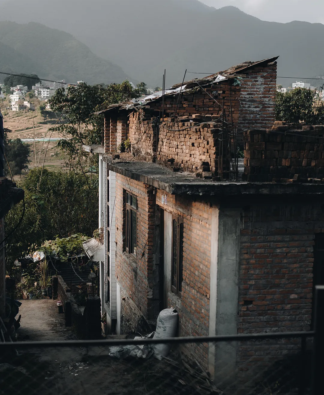Partially constructed brick building with sack near the door, surrounded by greenery and mountains in the background.