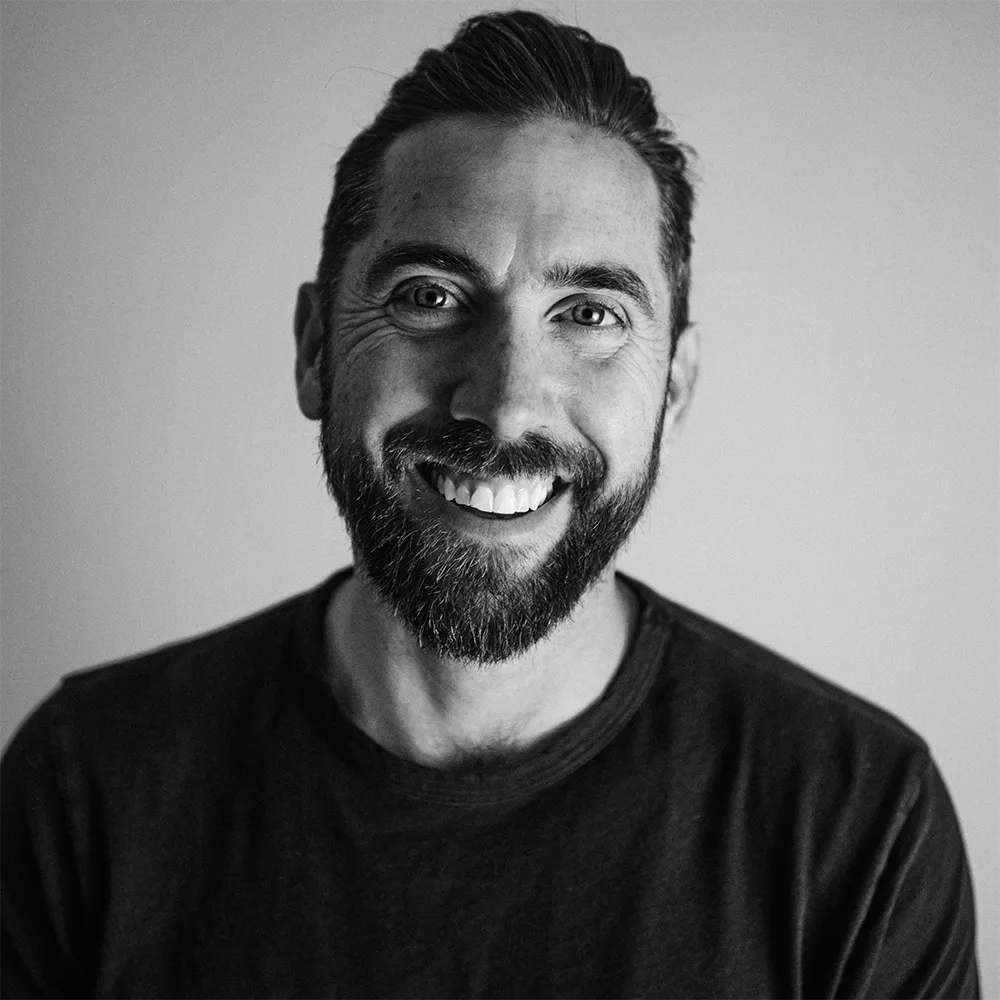 Smiling man with a beard and neatly styled hair wearing a dark shirt against a plain background.