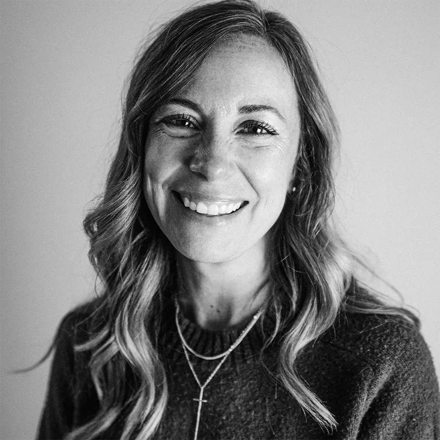 Black and white close-up portrait of a woman with long wavy hair smiling, wearing a dark sweater and layered necklaces.