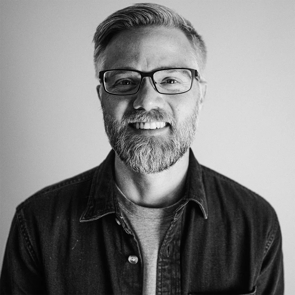 Portrait of a smiling middle-aged man with glasses, a beard, and wearing a dark button-up shirt over a t-shirt.