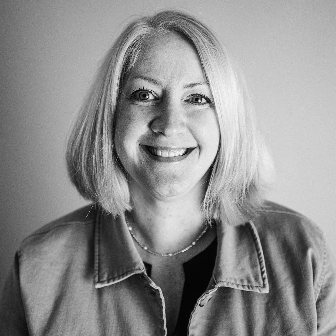 Smiling woman with shoulder-length blonde hair wearing a denim jacket and a beaded necklace.