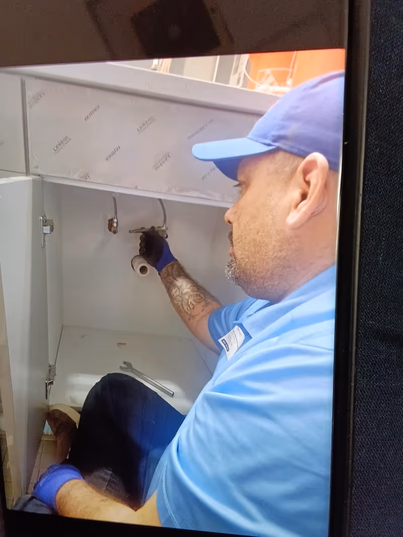 Plumber wearing a blue shirt and cap fixing pipes inside a kitchen cabinet.
