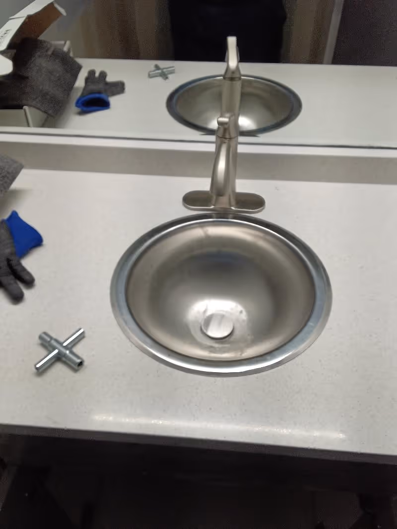 Bathroom countertop with a stainless steel sink and faucet, a pair of gloves, and a small metal cross-shaped tool.