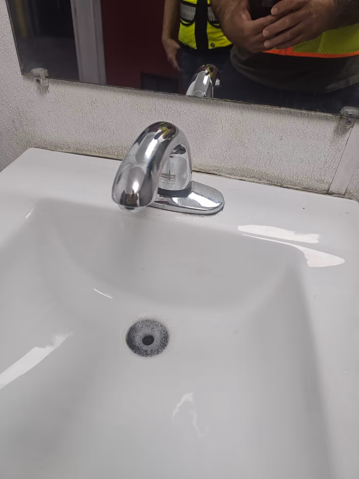White sink with a silver faucet mounted in front of a cracked and dirty mirror with a person partially reflected wearing a yellow safety vest.