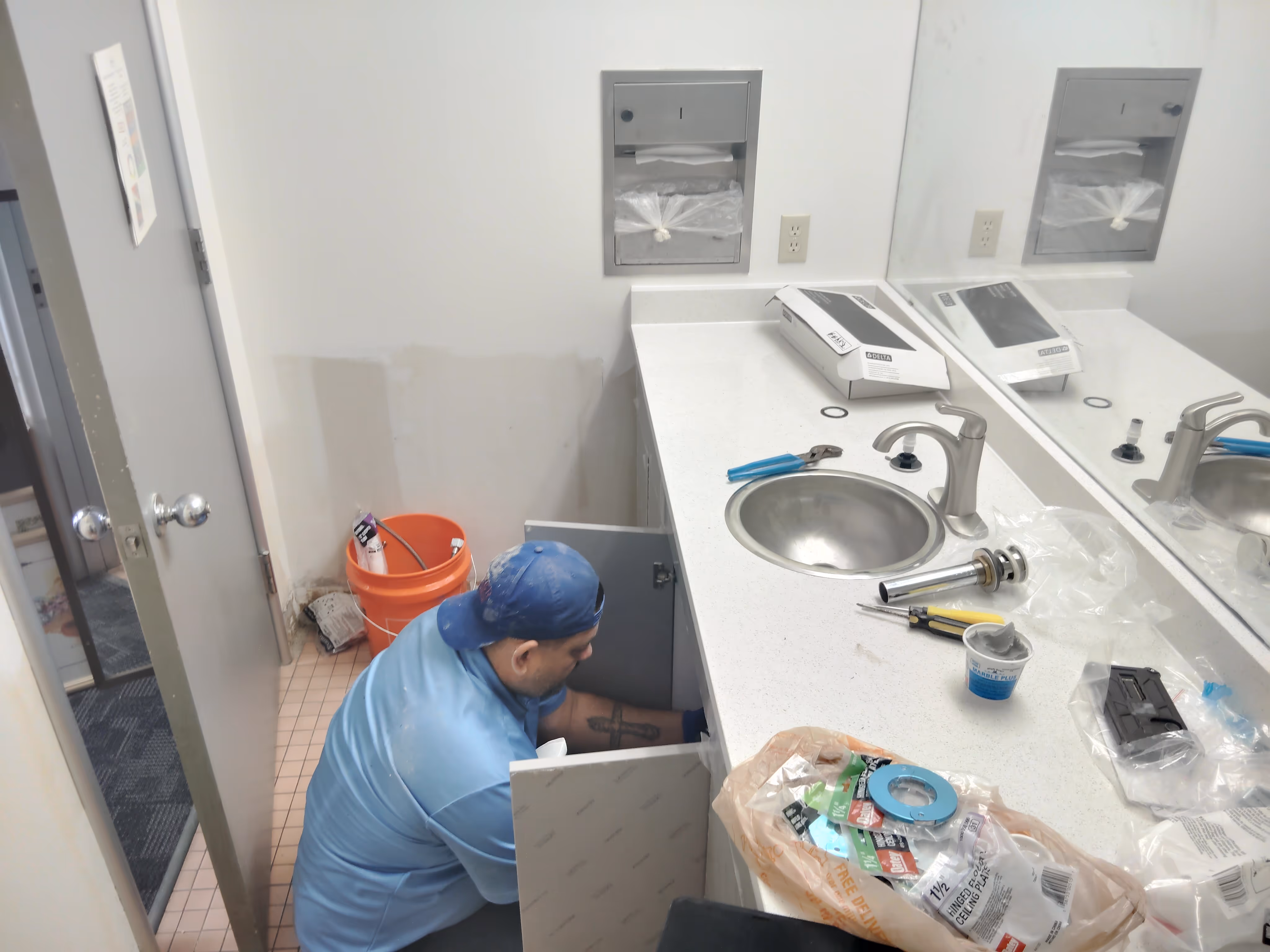 A man in a blue shirt and cap is working inside a bathroom cabinet under a sink, with plumbing tools and parts scattered on the countertop.
