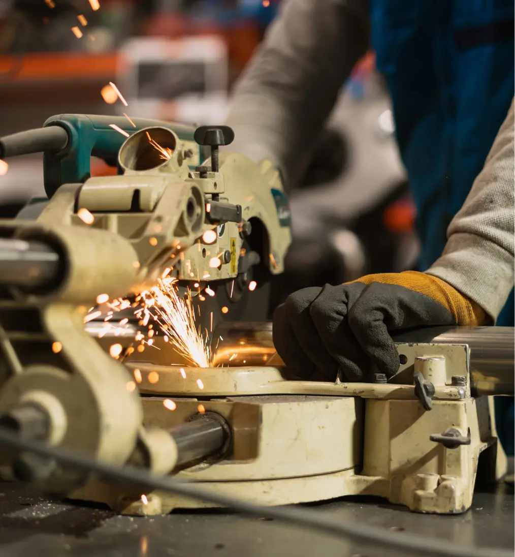 Worker wearing gloves operating a metal cutting saw with sparks flying in a workshop.