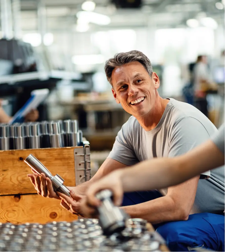 Smiling factory worker inspecting a metal part in a manufacturing environment.