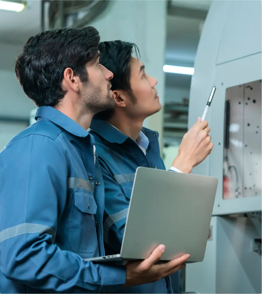 Two engineers in blue uniforms inspecting equipment, one holding a laptop and the other pointing with a marker.